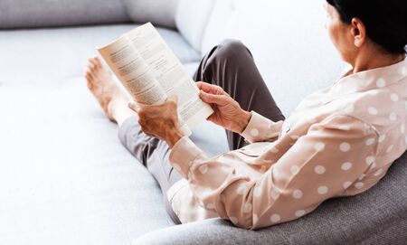 Middle-aged brunette woman on the gray sofa reading book, soft focus, stay at home concept, cozy backgroundの写真素材