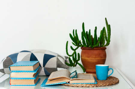 Cozy home interior decor: cup of coffee, stack of books, plants in pots, open book on a wicker stand, pillows on a white table. Distance home education.Quarantine concept of stay homeの写真素材