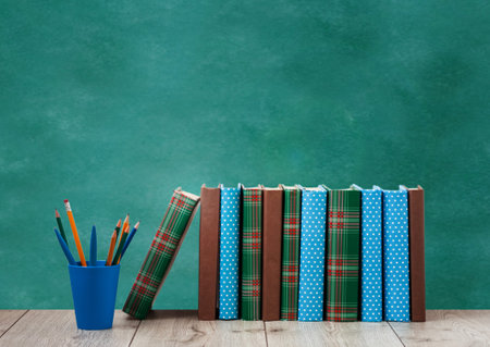 pile of books in colorful covers and pencils in holder on wooden table with green blackboard background. Distance home education. Back to school, quarantine concept of stay homeの写真素材