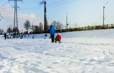 Children and their parents sledding from a snow slide in a city park with a frozen lake in Kiev,...