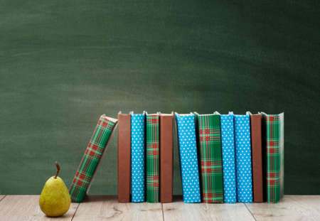 pile of books in colorful covers and pear on wooden table with green blackboard background. Distance home education. Back to school, quarantine concept of stay homeの写真素材