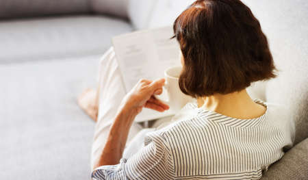 Middle-aged brunette woman with glasses on the gray sofa reading magazine with cup of coffee, soft focusの写真素材
