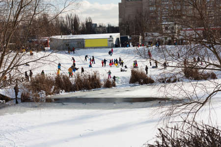Children and their parents sledding from a snow slide in a city park with a frozen lake in Kiev, Ukraineの写真素材