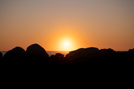 Sun setting on the ocean, behind a large rock in silhouetteの写真素材