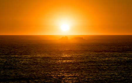 Vibrant orange sunset over ocean with reflections on water surface and rocks in silhouetteの写真素材