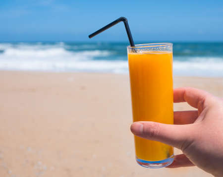 Hand holding glass of fresh orange juice at beach in summerの写真素材