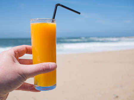 Hand holding glass of fresh orange juice on beach in summer