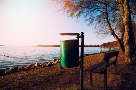 Wooden bench and green trash can near the lakeの写真素材