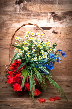 Cornflowers, chamomiles wheat and poppies in basket, wooden backgroundの写真素材