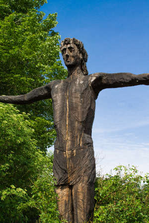 Side view picture of a wooden statue of Jesus who is standing in full pose with spread arms in front of a big tree and a blue skyのeditorial素材