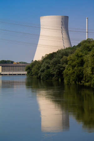The cooling tower of a nuclear power plant is reflected in a river. In front of it is a hydropower plant. Everything in close-up.の写真素材