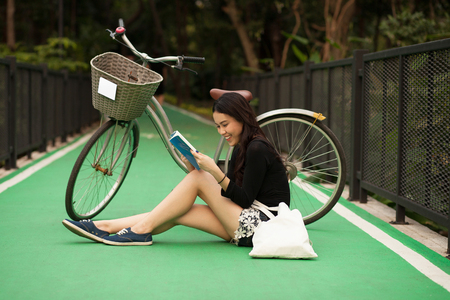 Pretty Thai girl reading book and sitting near by bicycle at the parkの写真素材
