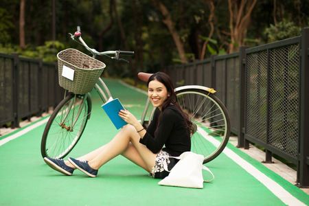 Pretty Thai girl reading book and sitting near by bicycle at the parkの写真素材