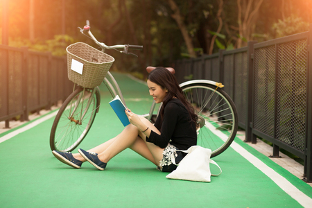 Pretty Thai girl reading book and sitting near by bicycle at the parkの写真素材