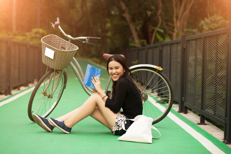 Pretty Thai girl reading book and sitting near by bicycle at the parkの写真素材