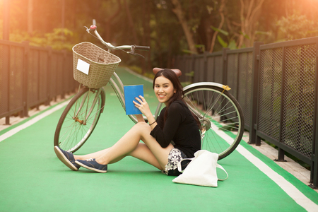 Pretty Thai girl reading book and sitting near by bicycle at the parkの写真素材