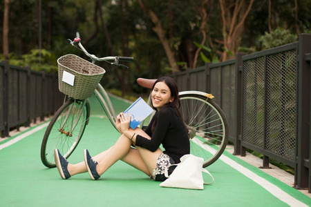 Pretty Thai girl reading book and sitting near by bicycle at the parkの写真素材