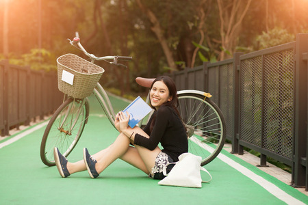 Pretty Thai girl reading book and sitting near by bicycle at the parkの写真素材
