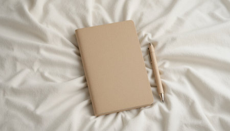 A blank notebook lies next to a pen on smooth white bedding, creating a calm and inviting workspace.の素材