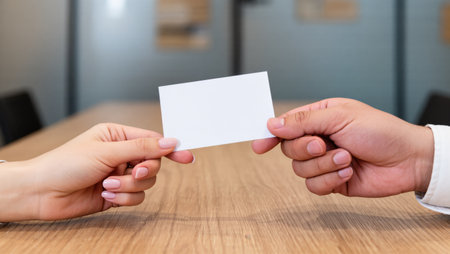 Two hands are exchanging a blank card over a wooden table in a contemporary office environment.の素材