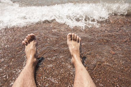 Closeup of a man's bare feet stand at wet on the beach , with a wave's edge foaming gently beneath themの写真素材
