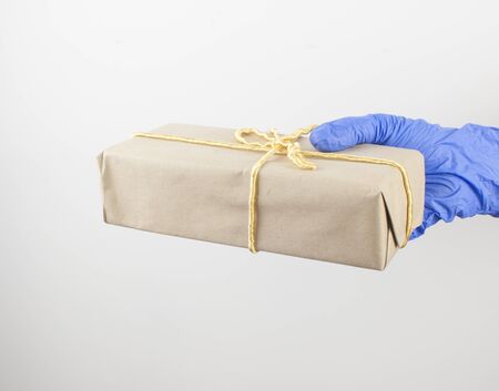a hand in a blue disposable glove holds gift box with a bow from on white wall background, copy...