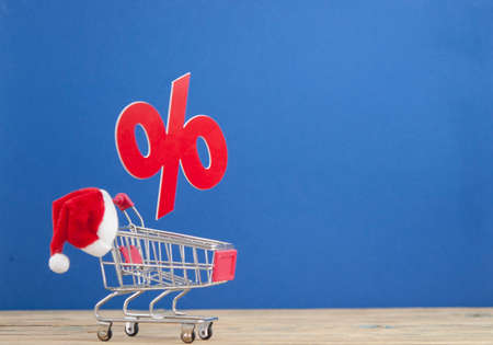 Christmas big sales: shopping cart with Santa hat and symbol of percent on wooden background