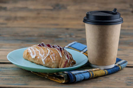 On a table made of natural wooden boards there is a ceramic plate with a sweet cherry bun and paper disposable coffee cup. Breakfastの写真素材