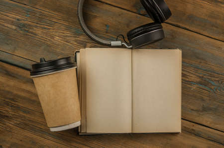 Open book with blank page, headphone and a cup of coffee in a disposable paper cup on a wooden...