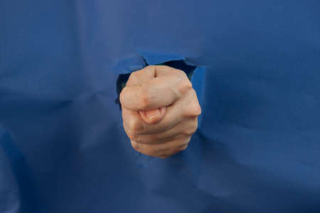 Female fist punching through blue paper background. War, struggle, conflict, feminist concept. Banner with copy space.の写真素材