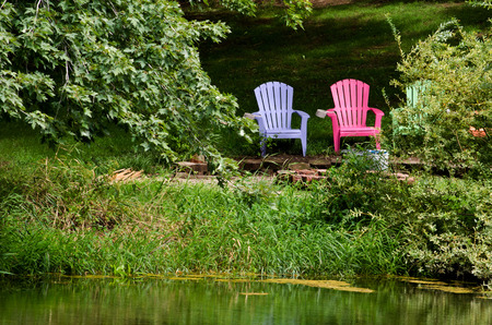 Two colorful adirondack chairs sit alongside a small pond waiting for someone to sit back and relaxの写真素材