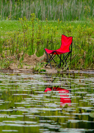 a red canvas chair sits beside a small calm lakeの写真素材