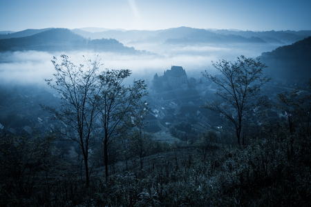 Mysterious misty morning over Biertan village, Transylvania, Romania. Blue colors. Halloween postcard concept.の写真素材