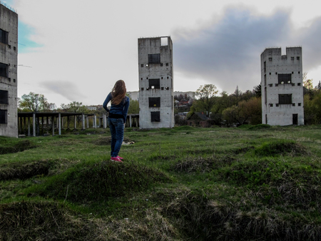 A young Slim girl with long blond hair stands on a vacant lot in front of three dilapidated towers on a cloudy day. She stands with her back on the hill and looks at the unfinished buildingsの写真素材