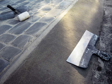 A large metal facade spatula lies on the surface of the concrete screed, and a white rubber mallet lies on the surface of the paving slabs. Construction work on site upgrades - outdoor flooringの写真素材