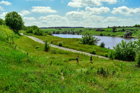 Two male tourists walk along a path among a spring-summer green meadow against a pond on a sunny...