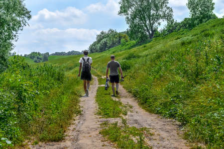 Two fishermen are walking along a meadow dirt road, view from the back. Male hikers with backpack and fishing rods among green nature on a sunny spring-summer dayの写真素材