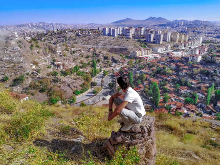 Adult young man squatting on a stone pillar and looking at the Ankara cityscape (Turkey) - view from...