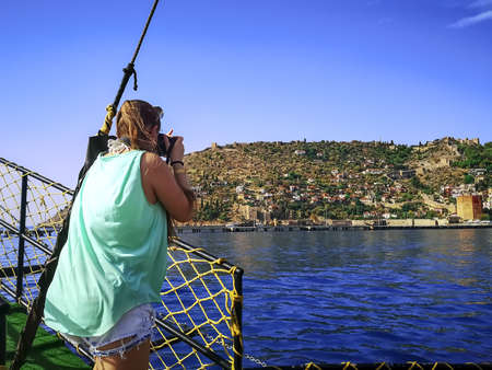 A young woman tourist photographs from the deck of a ship the peninsula with the Red Tower in Alanya...