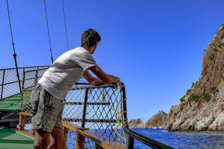 A tourist admires the rocks in the blue water from the ship. Man in summer casual clothes looks over the sea horizon while standing on the deck of the boat. The concept of sea tours in Alanya (Turkey)の写真素材