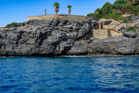 View from the sea to the abandoned park Emirgan Ulas Mesire Yeri in Alanya (Turkey). Two palm trees grow on a stone artificial terrace in a granite rock among turquoise waterのeditorial素材
