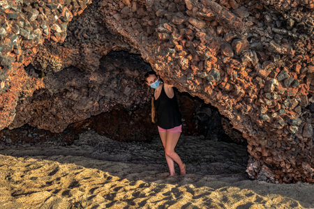 Young woman wearing a medical face mask peeks out of a cave at Cleopatra beach in Alanya (Turkey). Caucasian blonde tourist girl travels during the coronavirus epidemicの写真素材