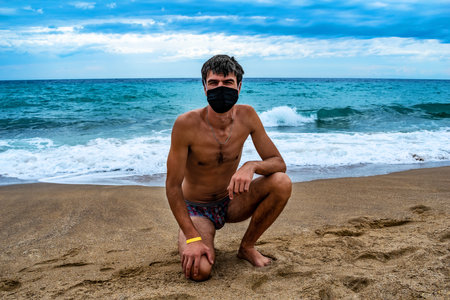 Cute young man sunbathing on beach wearing a medical face mask and swimming trunks. Adult guy sits on the sand against of a turquoise blue sea with waves. Travel concept during coronavirus quarantineの写真素材