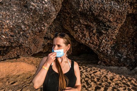 Young woman tourist straightens a medical mask on her face against the background of a cave on Cleopatra beach in Alanya (Turkey). Portrait of cute blonde caucasian girl outdoors on a summer sunny dayの写真素材