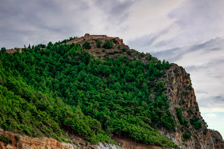A rock overgrown with coniferous forest with the ancient wall of the Alanya Castle (Turkey) at the top on a summer-autumn cloudy day. Beautiful mountain landscape with a medieval fortress among natureのeditorial素材