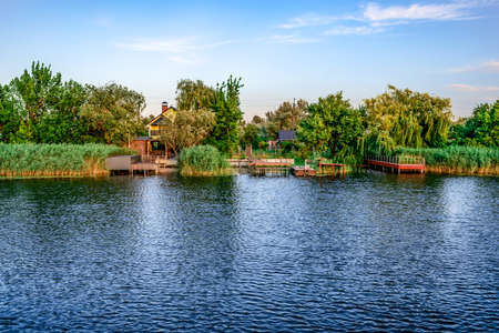 Residential houses with personal piers on Konka River coast in Kherson floodplains (Ukraine). Suburban area with buildings on the shore among green trees and reeds on the water surface backgroundの写真素材