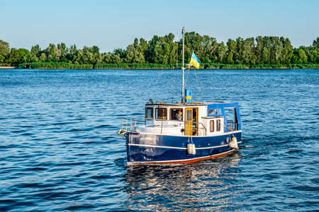 Boat on the Dnieper river in Kherson (Ukraine). Bright summer landscape of a small ship with a Ukrainian flag floating in blue waters against the backdrop of a green shoreのeditorial素材