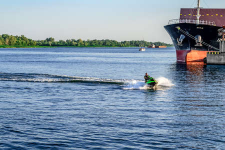 Kherson, Ukraine - July 22, 2020: A man on a water scooter glide past a large industrial ship in the Dnieper River in Kherson. Guy advertising water activities for tourists in a small Ukrainian townのeditorial素材