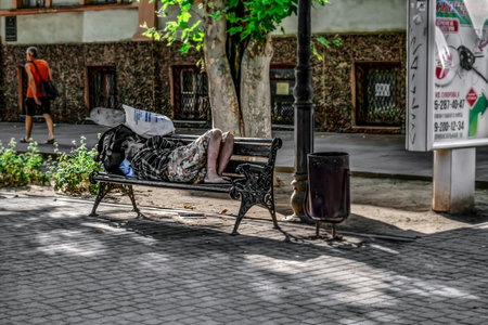 Kherson, Ukraine - July 22, 2020: A homeless man sleeps on a bench on Suvorova Street in Kherson. Barefoot man with dirty feet lies on a street bench on a summer sunny dayのeditorial素材