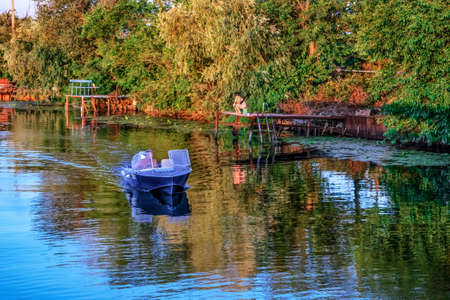 Kherson, Ukraine - July 22, 2020: People having a rest in nature on Konka River in Kherson plavny on a sunny summer evening. Woman fishing on the pier and a man sailing  on a boatのeditorial素材